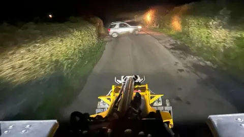 A view from what appears to be a combine harvester or some other farm machinery, driving along a narrow road seemingly lit only by the headlights, with the hedgerows either side blurred slightly from the movement. Ahead on the road is what appears to be a silver SUV, which by its position appears to have emerged from a gate to the left of the image.