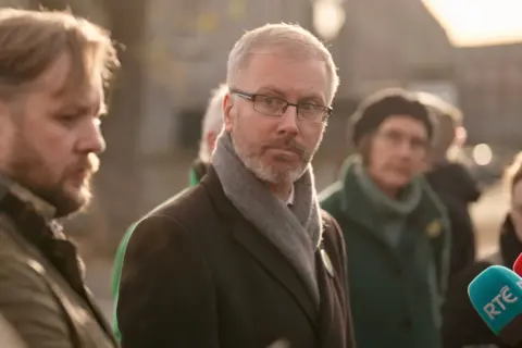 Roderic O’Gorman looks thoughtful portion    canvassing successful  the section  area, an RTE - Irish vigor   and TV - vigor   microphone visible. A antheral   and pistillate   either broadside  of the person   are besides  listening to the speaker, not pictured 