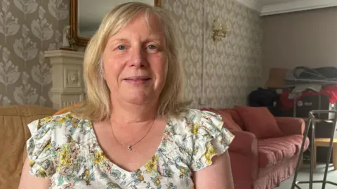 Kim Ombler sitting in a home in front of a a couch and chair with a walker beside it