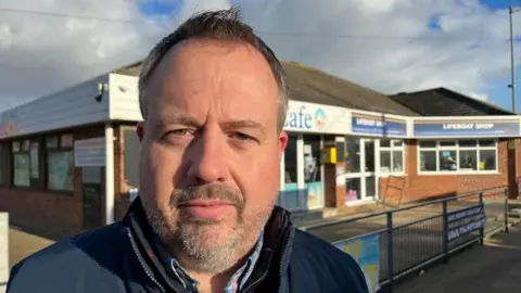 Andrew Turner/BBC A man with short brown hair that is slightly receding and a goatee beard. He is wearing a blue shirt, with a blue casual jacket over it. He is standing in front of the Beach Cafe. Next to it is the Hemsby Lifeboat Shop.