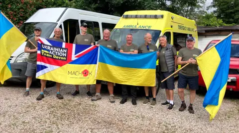A group of eight volunteers stand in a gravel parking area holding Ukrainian flags in front of a white minibus and a yellow ambulance. Several people wear matching “Ukrainian Action Team Devon” shirts. A large banner combining Ukrainian and Union Jack colours is displayed alongside a blue-and-yellow Ukrainian flag, with trees and parked vehicles in the background.

