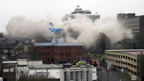 Corrine West Clouds of smoke arising from the demolition of a building which is hardly visible apart from a section covered by a blue sheet. There is a smaller brick building in front and a concrete car park to the right.