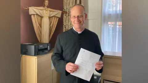 Supplied The Reverend Johannes Arens is standing holding a letter with a statue of Christ in the background.