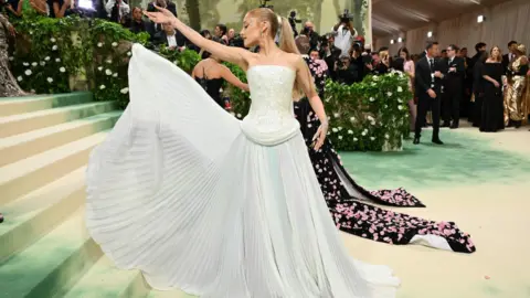 Getty Images Singer Ariana Grande is on the red carpet at the Met Gala 2024 in New York. She is wearing a floor-length, white ballgown and is showing off the pleats on the bottom. She has her head turned to the side and her long, blonde hair is tied back in a ponytail.