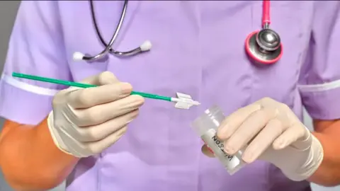 A female nurse holds a green specimen stick close to a jar while wearing gloves. She has a purple uniform on with a stethoscope around her neck.