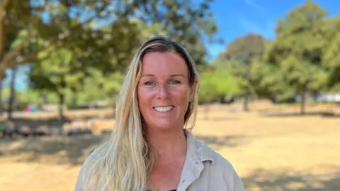 Rebecca Coley está de pie en un espacio verde abierto en un día soleado. Lleva una sobrecamisa o chaqueta de color beige claro sobre una blusa oscura abotonada. Al fondo se aprecian árboles maduros con ramas extendidas, hierba seca y zonas de sombra en el suelo. El cielo es despejado y de un azul brillante, y el fondo está ligeramente desenfocado, lo que centra la atención en la persona en primer plano.