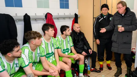 Tunley Athletic FC Harry Redknapp in a changing room talking to several footballers sitting on a bench, wearing their green and white striped Specsavers kit