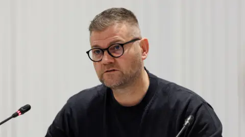 PA Media Colin is sat down in front of two small microphones. Ge is wearing a black t-shirt and dark round-rimmed glasses. His fair is greying and short and he has a short grey-speckled stumble. 