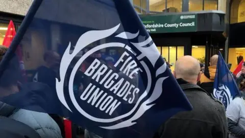 protestors have gathered outside oxfordshire county councils officers waving blue flags with "fire brigades union" written on them