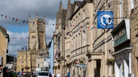 A street view of Cirencester. There are stone buildings with bunting hanging across the street. There is a church in the background.