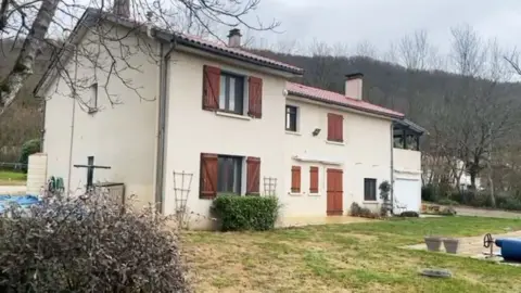 White house with brown shutters and windows with green meadows, there are trees and hills in the background