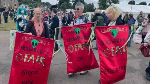 Three people are standing on a walkway in a field holding bright red banners that read "Strawberry Fair". There are several people in the background, walking.