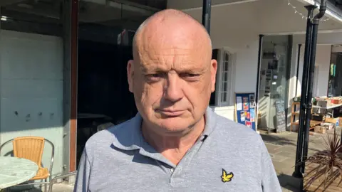 Malcolm Pollard is standing in a street arcade slightly smiling at the camera. He has a shaven head and is wearing a grey T-shirt with buttons on the neckline. Behind him is a table and chair and further behind him is a table filled with boxes of books. It is sunny.