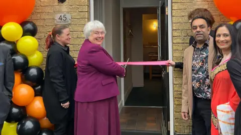 A councillor wearing a purple co-ord made up of a purple blazer and purple skirt. She is cutting a ribbon ahead of the opening of the restaurant. 
The owners are standing on the side smiling at the camera. The woman is wearing a traditional Indian dress called a saree. The man is wearing a flowered shirt and a brown blazer. There are balloons by the side of the door. 