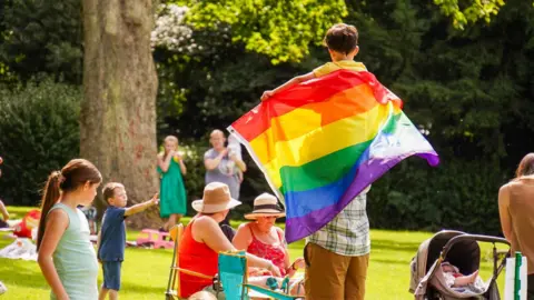 Families celebrating at a Pride event in Gateshead's Saltwell Park. A boy sits on a man's shoulders. The boy has the rainbow Pride flag draped over his shoulders.