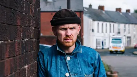 Rory Carson Rory Carson in a blue denim jacket with the collar turned up. He also wears a gray cap. He is looking directly at the camera with a serious expression as he leans against a red brick wall in a street. There is an ice cream van in the background