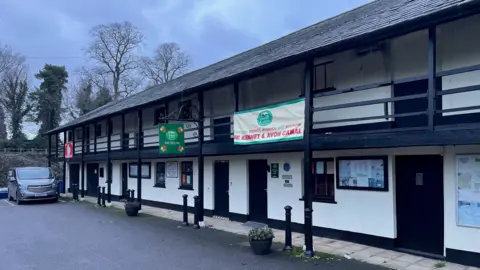 The front of the Kennet and Avon Canal HQ, which is a long white building with black trim and grey roof. It has short black pillars in front of it and there are trees to the left of the building as you look at it. There is a grey can parked to the left hand side. The sky is cloudy.