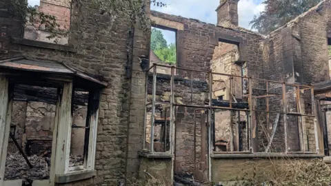 The ruin of Holly Lodge after it was gutted by fire. There is no roof or windows and the two-storey stone building is an empty shell with signs of extensive fire damage. The branches of trees and other plants surround the property.