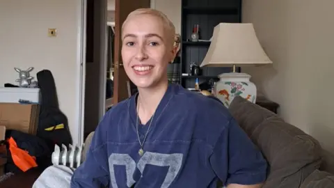 A woman with short blonde hair, wearing a blue top with a pendant around her neck. She is sat in a living room. 