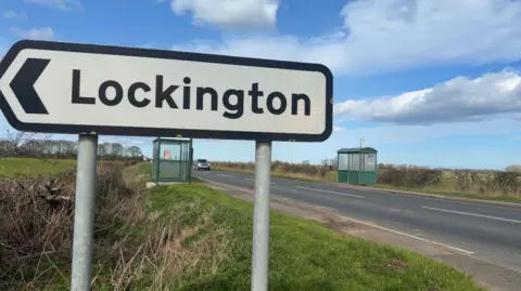 Jasmine Lowe/BBC News A road sign pointing left to Lockington. Behind it is a busy A road with two green bus shelters opposite each other on either side of the road. 