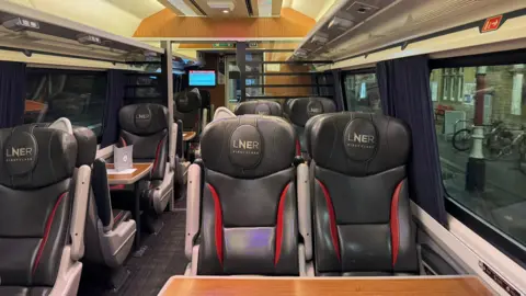 225 Preservation CIC Inside one of the former LNER carriages. Passenger seats line either side of the carriage with tables in front of them. There is also LNER branding on the headrests of the seats. 