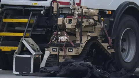 PA Media A bomb disposal robot positioned on a paved surface next to a large vehicle, which appears to be a specialized truck. The robot is tan-coloured and equipped with tracks for movement, as well as an articulated arm for handling objects. In front of the robot, there is a black protective suit lying on the ground, likely a bomb disposal suit, along with a sturdy black case and a green bag resting on top of it. The background includes part of the truck’s side, visible wheels, and steps leading up to the cab.