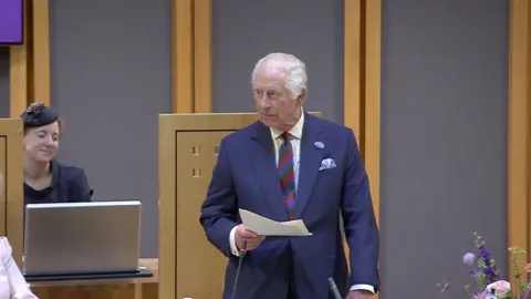 The King speaking from the dais in the Senedd chamber, in a blue suit and holding a paper in his right hand