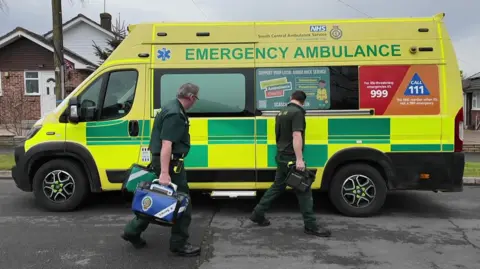 Two male paramedics walking on a residential road outside a yellow and green NHS ambulance. They are wearing green paramedic uniforms and carrying medical bags. In the background is a bungalow. The ambulance has "emergency ambulance" written on it.