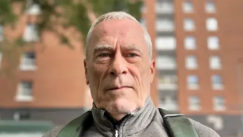  Resident Mark Barrett looking into the camera with a tower block, not in focus, behind him in the background