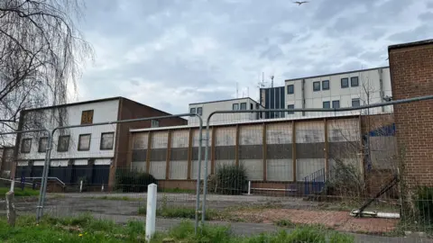 Exterior of a disused building. A metal barrier blocks entrance to the site which is boarded up. The building is large and spans across a wide area. It is white with red bricks.