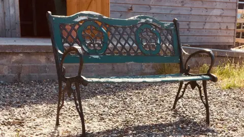 Northern Men in Sheds A decorative metal bench with turquoise paint that is peeling off the back rest. It is placed on some gravel, with a shed in the background.