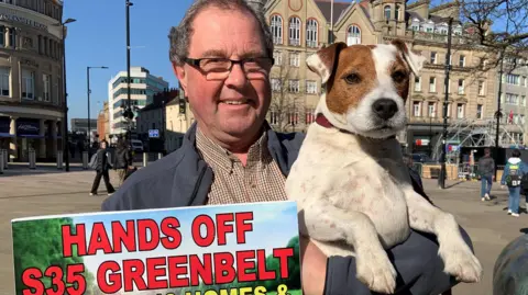 A man with short dark hair and glasses is wearing a brown checked shirt and a blue jacket. His is holding a brown and white short-haired terrier dog and holding a colourful placard which says Hands Off S35 Green belt