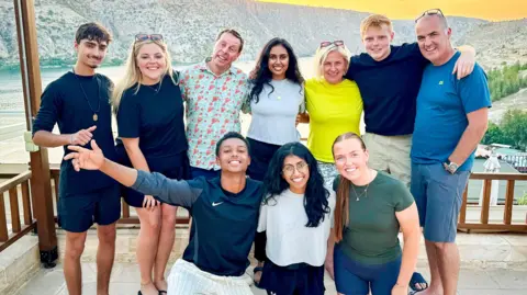 BBC/Studio Lambert Kush, Katie, Mark, Jo, Puja, Roshni, Margo, Molly, Harrison, Andrew pose as a group on a balcony in Halfeti, Turkey with a river and mountain range behind.
