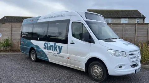 White minibus van parked on gravel in front of a big fence. It has Slinky branding on the side. 