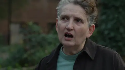 A middle-aged woman with short, greying curly hair and a brown shirt