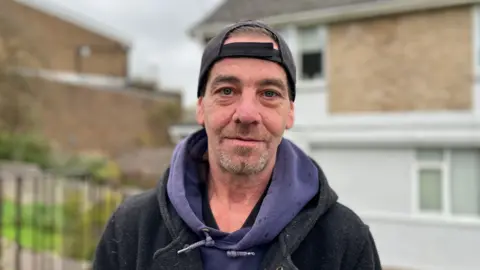A man is facing the camera, he's got brown/grey stubble and blue eyes. He's wearing a black baseball cap back-to-front, a blue hoodie and a black coat. Behind him, out of focus, is a house