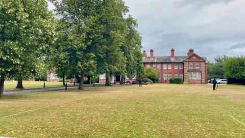 An ornate building with a large field in front of it as well as a playing field and trees. There is a row of trees to the left hand side of the image, obscuring part of the building. Two people in black clothing are standing in front of the building.