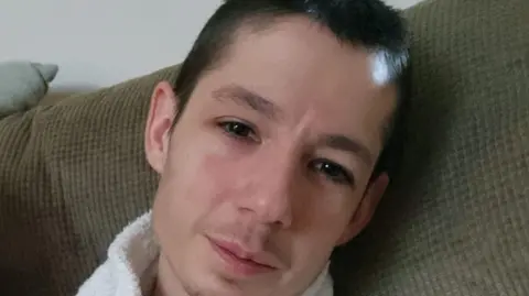 Bradley Roberts, sitting on a sofa, looking straight at the camera. He has short dark hair and a light stubble above his lips. He is wearing a white top. The sofa is a light green colour. You can see a wall behind him. 