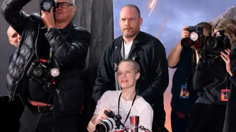 Liz Hatton holding a camera on the red carpet, surrounded by fellow photographers. 