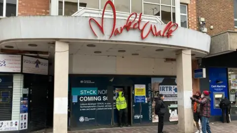 The outside of the former Market Walk shopping centre, with a man in a high-vis jacket in the doorway, and three other people to the right. A man is by a cash machine to the right. There is a white frame round the building the front boarded up with signs on it. 