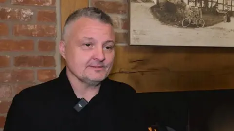 A man stands in front of a fire place. He has short grey and black hair and a black shirt. Behind him is a wooden mantle piece and a red brick wall. The flames from the fire are just visible at the bottom of the image.