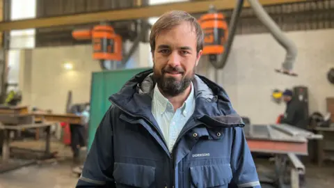 A man in his mid thirties with a beard wearing a blue shirt and thick blue jacket. He is standing in a factory which is out of focus.