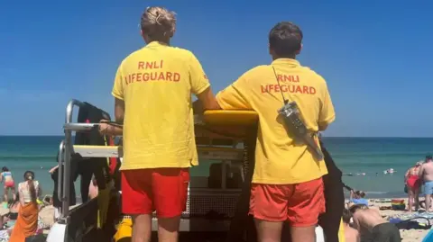 Two lifeguards, wearing yellow tops which said 'RNLI Lifeguard' and red shorts. There are looking out to the water and facing away from the camera. There are people on the beach facing the sea. In the background is the blue sea and light blue sky. 
