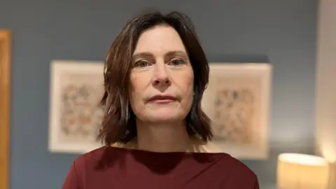 Bernie has brown shoulder length hair and is wearing a burgundy coloured top. She is looking directly at the camera. The wall behind her is blue and there is a lamp in the background