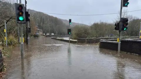 West Yorkshire Police A heavily flooded road with no cars on it. Traffic lights, which are on green, flank the road. The sky is grey and the trees in the background are bare and autumnal.