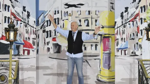 Getty Images Paul Costelloe is dressed in a waistcoat with a black tie, blue shirt and jeans. A bright cartoon backdrop showing buildings is behind him.