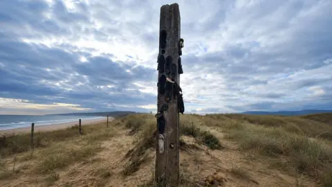 Jayne Gray A large wooden post has been buried upright at the top of a sand dune. Shoe soles have been nailed to the post up the sides. Grass is covering the dunes with the beach and sea to the left.