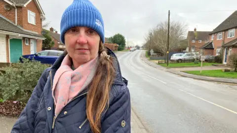 Henry Godfrey-Evans/BBC A woman with a blue woollen hat, a pink scarf and a blue coat with a long road stretching off into the distance