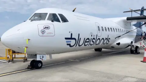 A Blue Islands aircraft parked on an airport tarmac under a partly cloudy sky. The white plane features the 'blueislands' logo on its side, with yellow and red safety markings near the nose wheel and a stairway positioned at the rear door.