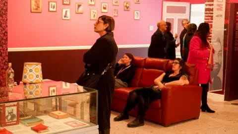 Ayesha Jones A group of people walk through an art gallery. One woman stands with her arms crossed looking at a wall. Two others are sat on a large sofa behind her.